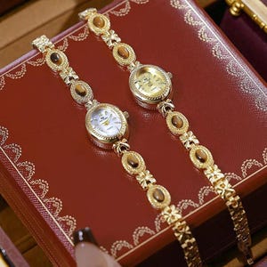 Puede incluir: Dos relojes de pulsera dorados con esferas ovaladas y correas decorativas. Las esferas son blancas y doradas, con números romanos. Las correas presentan piedras marrones ovaladas y detalles florales. Los relojes se exhiben sobre una superficie roja.