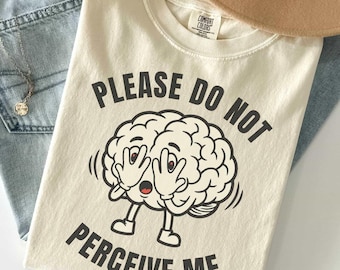 Peut inclure: Un t-shirt couleur crème avec une illustration de cerveau en dessin animé se couvrant les yeux avec ses mains. Le texte "PLEASE DO NOT PERCEIVE ME" est imprimé en lettres capitales noires au-dessus et en dessous du dessin. Col rond.