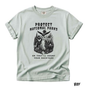 May include: A light green t-shirt with rolled-up sleeves. The front features a black graphic with the text "PROTECT NATIONAL PARKS" above an illustration of a Bigfoot, a ghost, and other forest creatures. Below the graphic is the text "OR THEY'LL HAUNT YOUR BACKYARD."