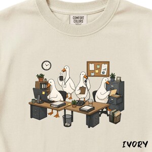 May include: Ivory-colored sweatshirt featuring a whimsical illustration of four geese working in an office setting. The geese are depicted at desks with computers, phones, and coffee cups. The word "IVORY" is printed at the bottom.
