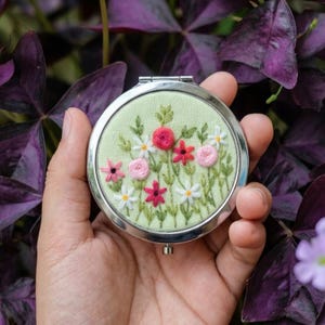Op de afbeelding: Een ronde, zilveren compacte spiegel met een geborduurd bloemenontwerp. De borduursels tonen roze, rode en witte bloemen met groene bladeren op een lichtgroene achtergrond. De spiegel wordt in een hand gehouden tegen een achtergrond van paarse bladeren.