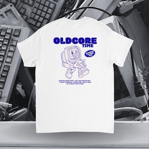 Peut inclure: T-shirt blanc avec un dessin bleu d'un personnage d'ordinateur rétro et les mots "OLDCORE TIME". Le t-shirt comporte également le texte "BE PATIENT I'M FROM 1990" et une citation sur la patience. Des claviers d'ordinateur anciens sont en arrière-plan.