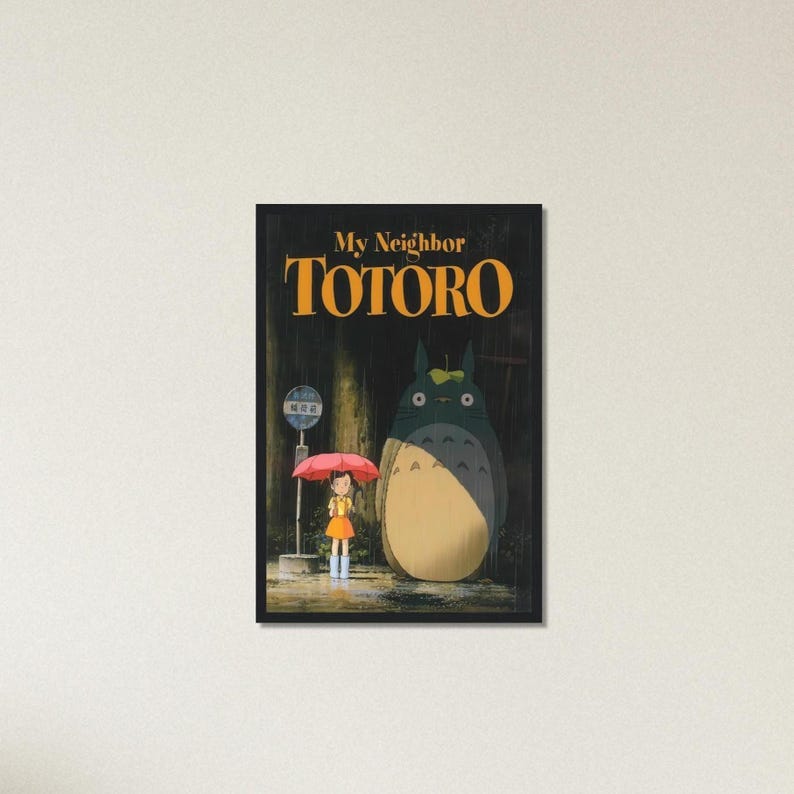 Pode incluir: Um poster emoldurado do filme de anima&ccedil;&atilde;o "Meu Vizinho Totoro". A imagem mostra uma menina com um guarda-chuva vermelho em um ponto de &ocirc;nibus, junto com uma criatura grande e peluda. O t&iacute;tulo "TOTORO" est&aacute; em letras laranja.