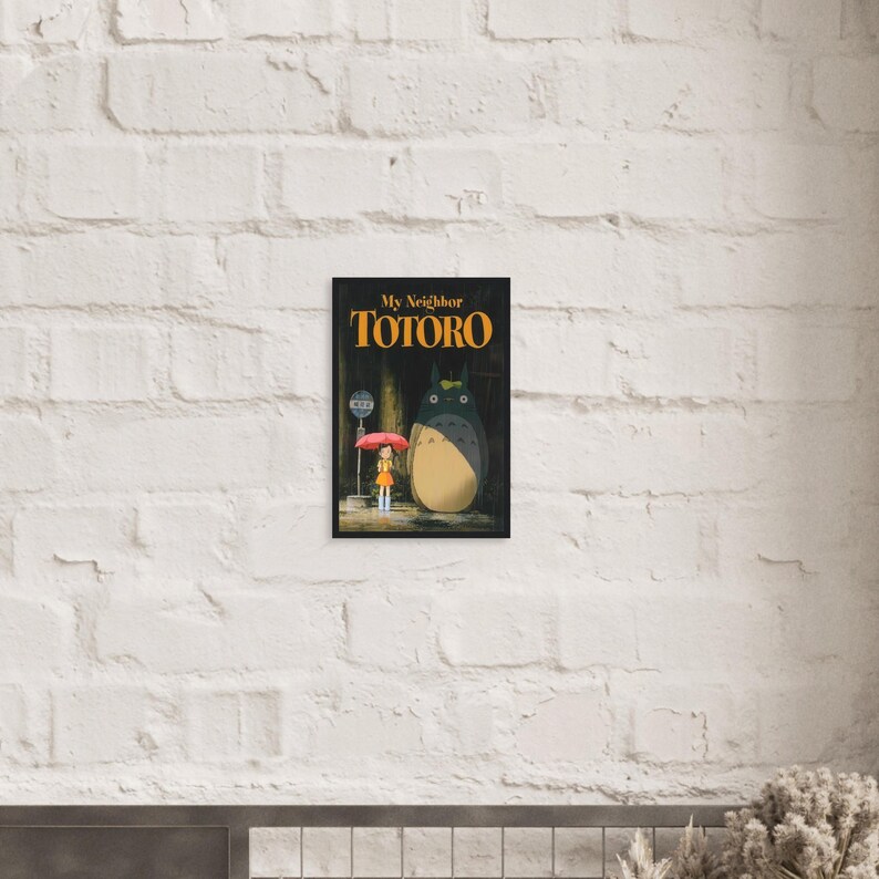 Pode incluir: Um poster emoldurado com o filme de anima&ccedil;&atilde;o "Meu Vizinho Totoro". A obra de arte retrata uma menina com um guarda-chuva vermelho em um ponto de &ocirc;nibus com Totoro. O t&iacute;tulo est&aacute; em letras laranja sobre um fundo preto.