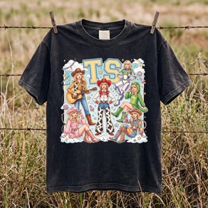 K&ouml;nnte beinhalten: Ein schwarzes Kurzarm-T-Shirt mit einem Grafikdruck von sechs illustrierten Figuren in verschiedenen Outfits, darunter ein Cowgirl mit Gitarre, ein Raumanzug und ein gr&uuml;nes Dinosaurierkost&uuml;m, mit den gro&szlig;en gelben Buchstaben TS dar&uuml;ber.
