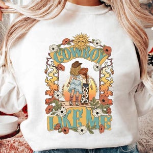 May include: A white crewneck sweatshirt featuring a graphic print of two figures in cowboy hats and boots standing together. The design includes the text "COWBOY LIKE ME" in blue, surrounded by orange flames, flowers, and a sun illustration.