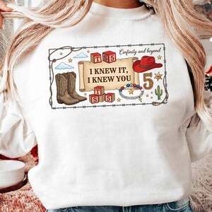 May include: A white sweatshirt featuring a graphic print of a scroll with the text I KNEW IT, I KNEW YOU, surrounded by cowboy boots, a red hat, alphabet blocks with TS5, a beaded bracelet, a cactus, and the text Cinfinity and beyond.