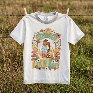 May include: A light gray short-sleeve t-shirt featuring a graphic print of two figures in cowboy hats and boots standing before a sunset. The design includes floral accents, flame motifs, and the text "COWBOY LIKE ME" in a bold, retro-style font.