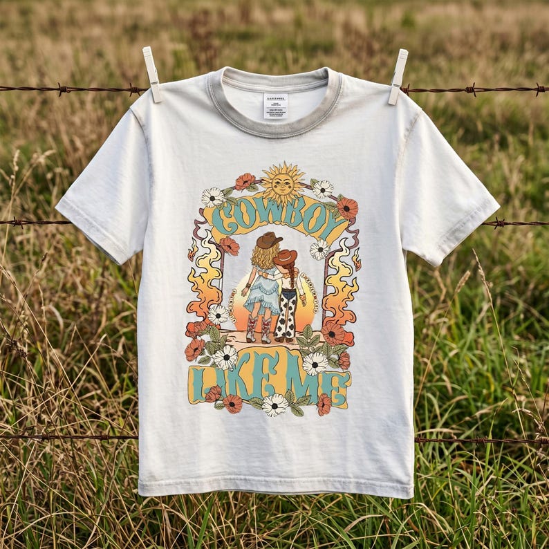 May include: A light gray short-sleeve t-shirt featuring a graphic print of two figures in cowboy hats and boots standing before a sunset. The design includes floral accents, flame motifs, and the text "COWBOY LIKE ME" in a bold, retro-style font.
