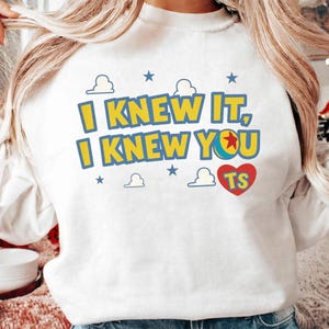 May include: A white long-sleeve sweatshirt featuring a graphic print with the text I KNEW IT, I KNEW YOU in yellow letters with blue outlines. The design includes a colorful ball, small blue stars, white cloud outlines, and a red heart with TS.
