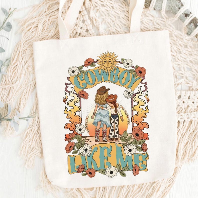 May include: A white canvas tote bag featuring a colorful graphic print of two figures in cowboy hats and boots standing together. The design includes a sun, flowers, flames, and the text "COWBOY LIKE ME" in a bold, retro-style font.