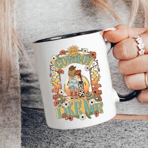 May include: A white ceramic mug with a black handle and rim, featuring a graphic illustration of two figures in cowboy hats embracing. The design includes floral accents, a sun, and the text "COWBOY LIKE ME" in a bold, retro-style font.