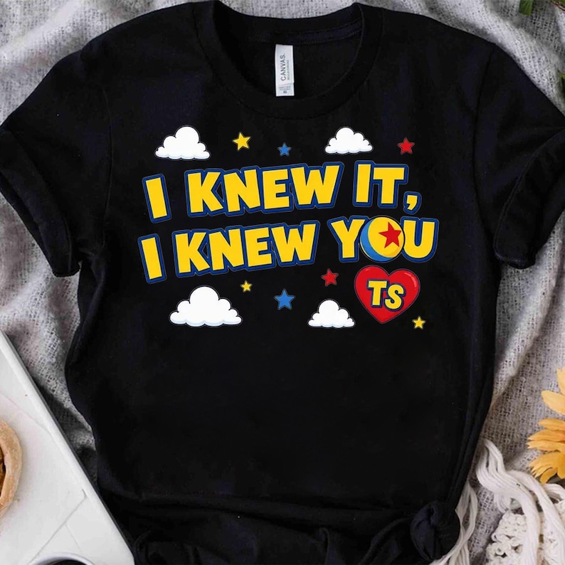 Peut inclure: Un t-shirt noir &agrave; manches courtes avec le texte jaune I KNEW IT, I KNEW YOU, un c&oelig;ur rouge contenant les lettres TS et un motif de balle. Le design comprend des nuages blancs et de petites &eacute;toiles rouges, bleues et jaunes sur le devant.