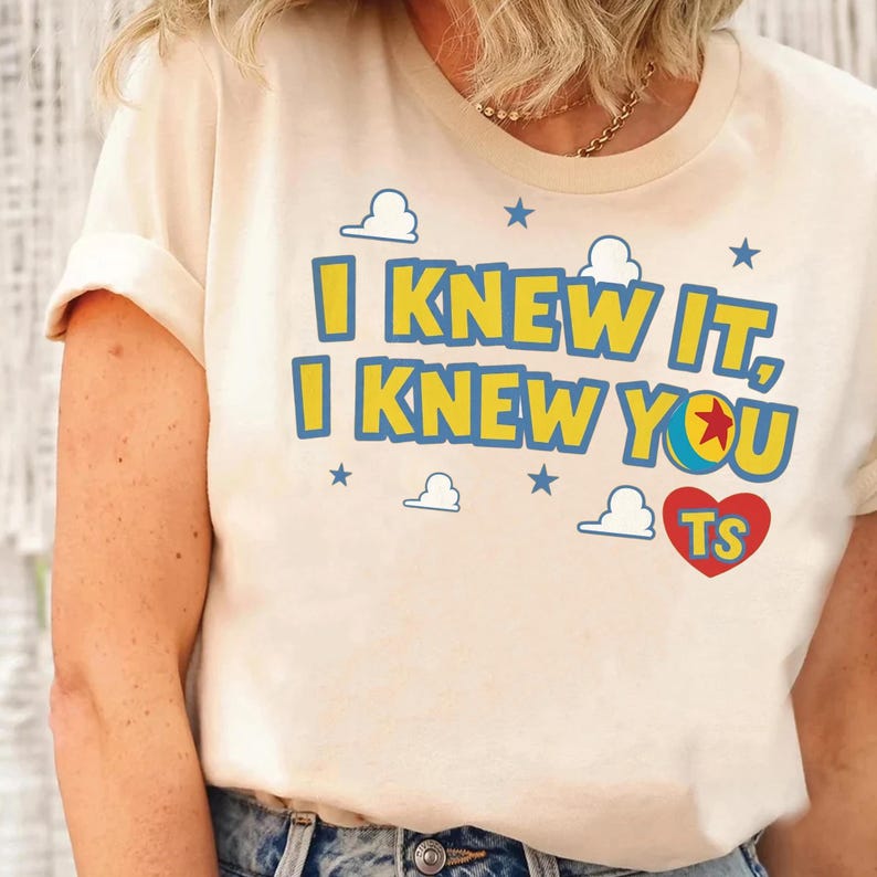 May include: A cream-colored t-shirt featuring the yellow text I KNEW IT, I KNEW YOU with blue outlines. The design includes a colorful ball icon, blue stars, white cloud shapes, and a red heart containing the yellow letters TS.