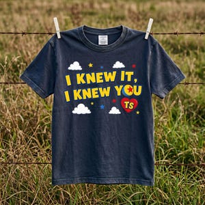 Peut inclure: Un t-shirt gris fonc&eacute; &agrave; manches courtes avec le texte jaune I KNEW IT, I KNEW YOU, un graphique de balle color&eacute;e, un c&oelig;ur rouge avec les lettres TS, ainsi que des nuages blancs et de petites &eacute;toiles imprim&eacute;s sur la poitrine.