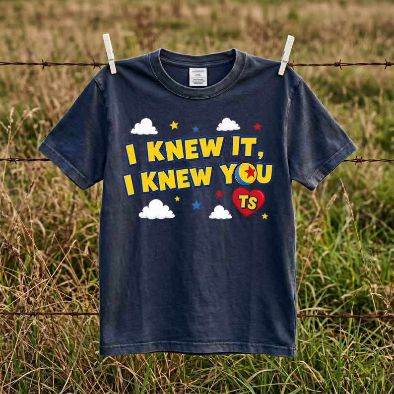 Peut inclure: Un t-shirt gris fonc&eacute; &agrave; manches courtes avec le texte jaune I KNEW IT, I KNEW YOU, un graphique de balle color&eacute;e, un c&oelig;ur rouge avec les lettres TS, ainsi que des nuages blancs et de petites &eacute;toiles imprim&eacute;s sur la poitrine.