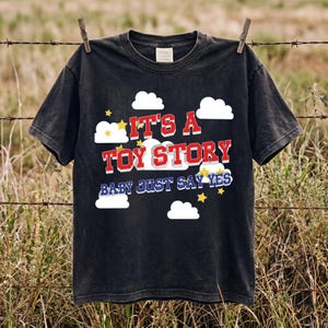 May include: A black short-sleeved t-shirt featuring a graphic print of white clouds and yellow stars. The text reads IT'S A TOY STORY in red block letters with a white outline, followed by the blue text BABY JUST SAY YES in a bold font.