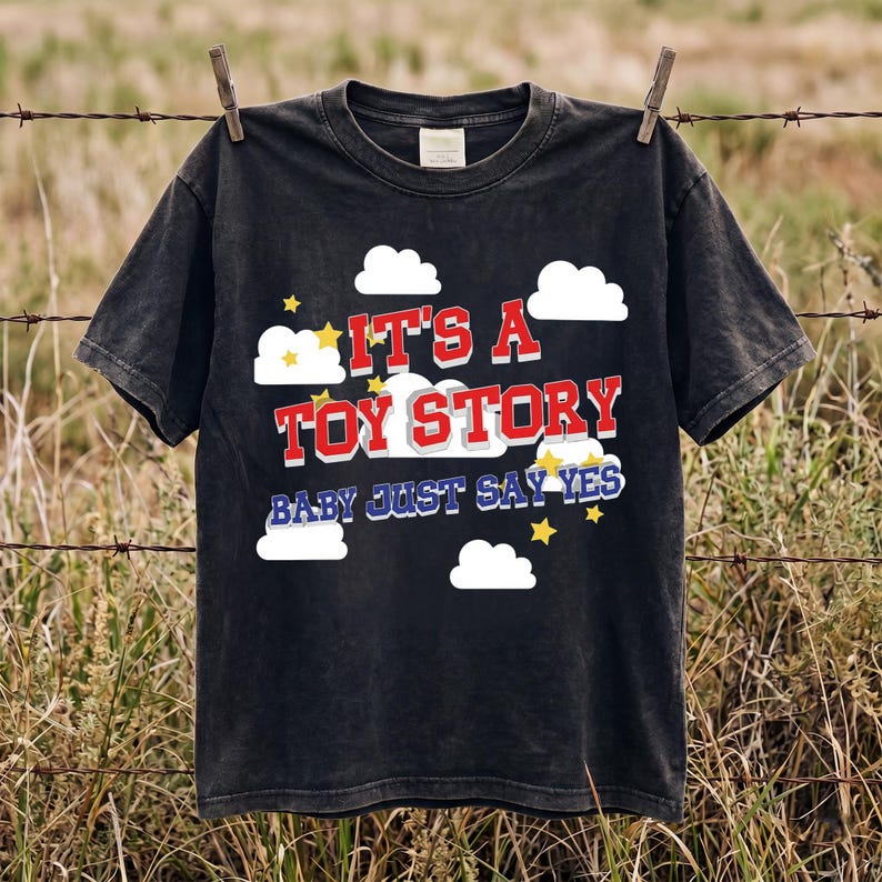 May include: A black short-sleeved t-shirt featuring a graphic print of white clouds and yellow stars. The text reads IT'S A TOY STORY in red block letters with a white outline, followed by the blue text BABY JUST SAY YES in a bold font.