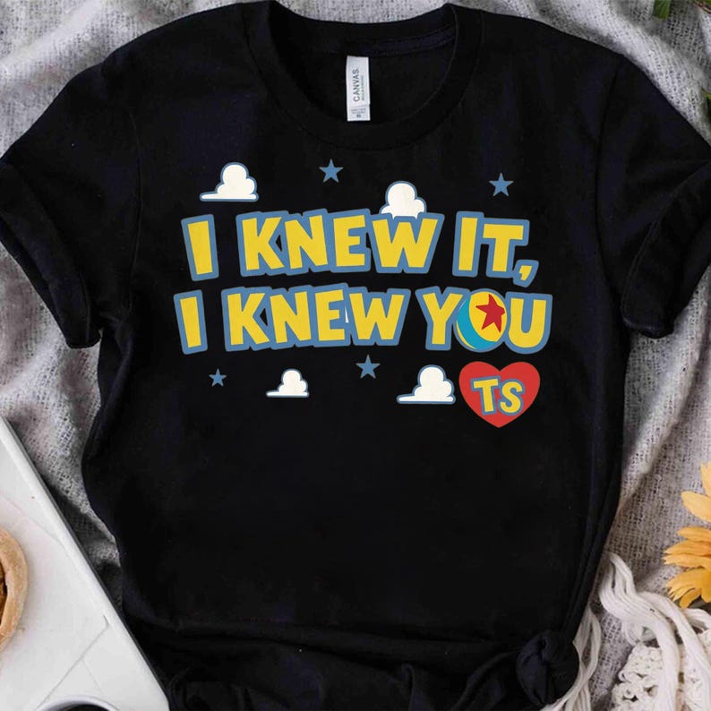 May include: A black short-sleeved t-shirt featuring the yellow text I KNEW IT, I KNEW YOU with a colorful ball graphic, a red heart containing the letters TS, and small white cloud and blue star icons printed on the front chest area.