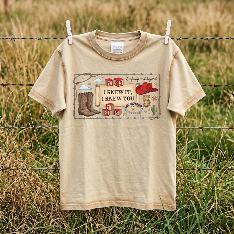 May include: A beige short-sleeve t-shirt featuring a graphic print with the text "I KNEW IT, I KNEW YOU" and "Cinfinity and beyond". The design includes illustrations of cowboy boots, a red hat, alphabet blocks, a bracelet, and a cactus.