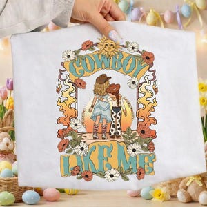 May include: A white t-shirt featuring a graphic print of two figures in cowboy hats standing together. The design includes a sun, flowers, and the text COWBOY LIKE ME in a retro font. The figures wear a dress and cow-print pants respectively.