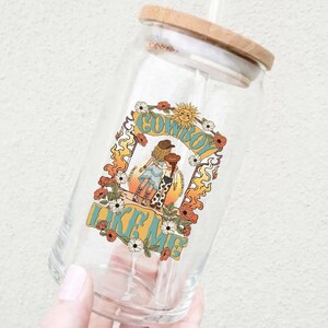 May include: A clear glass tumbler with a bamboo lid and a straw, featuring a colorful graphic print of two figures in cowboy hats, surrounded by flowers, flames, and a sun, with the text COWBOY LIKE ME printed in bold blue letters.