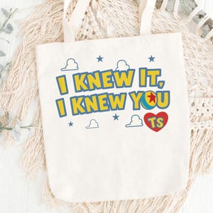 May include: A cream-colored canvas tote bag featuring the text I KNEW IT, I KNEW YOU in yellow with blue outlines. The design includes a colorful ball icon, small blue stars, white cloud outlines, and a red heart containing the letters TS.