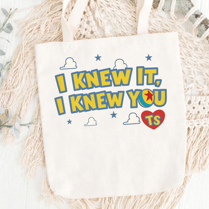 May include: A cream-colored canvas tote bag featuring the text I KNEW IT, I KNEW YOU in yellow with blue outlines. The design includes a colorful ball icon, small blue stars, white cloud outlines, and a red heart containing the letters TS.