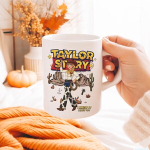 May include: A white ceramic mug featuring a colorful graphic of a cowgirl character in a desert landscape. The text reads TAYLOR STORY at the top and I KNEW IT I KNEW YOU at the bottom. The design includes cacti, rock formations, and stars.