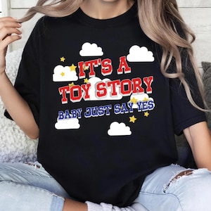 May include: A black t-shirt featuring a graphic print with white clouds and yellow stars. The text reads IT'S A TOY STORY in red block letters with a white outline, followed by the blue text BABY JUST SAY YES with a white outline.