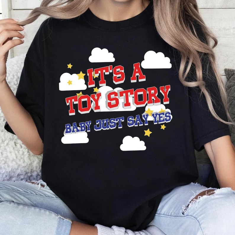 May include: A black t-shirt featuring a graphic print with white clouds and yellow stars. The text reads IT'S A TOY STORY in red block letters with a white outline, followed by the blue text BABY JUST SAY YES with a white outline.
