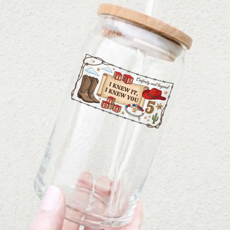 May include: A clear glass tumbler with a bamboo lid and a graphic print featuring brown cowboy boots, red cowboy hat, alphabet blocks with the text TS5, a beaded bracelet, a cactus, and a scroll with the text I KNEW IT, I KNEW YOU.