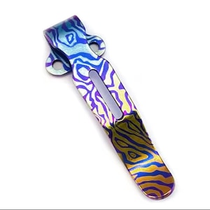 May include: A multi-colored pocket clip with a unique pattern. The clip features a vibrant mix of blue, purple, and gold hues, creating a striking visual effect. The design includes a curved top with two holes and a long, slender body with a central cutout.