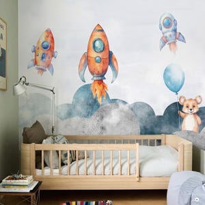 May include: A child's bedroom with a light wood cot and a wall mural featuring watercolour rockets, clouds, and a bear holding a blue balloon. The rockets are orange, blue, and white, with stylized flames.