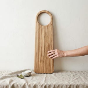May include: A rectangular wooden cutting board with a circular handle. The board is made of light brown wood with visible grain. A small bowl of salt and a sprig of lavender are on a linen surface.