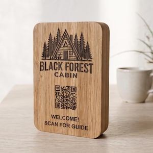 Puede incluir: Un letrero rectangular de madera marrón claro con un grabado láser de una cabaña en un bosque. El texto dice BLACK FOREST CABIN, seguido de un código QR y el texto WELCOME! SCAN FOR GUIDE en letras de color marrón oscuro.