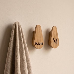 Könnte beinhalten: Zwei hellbraune Wandhaken aus Holz in Tropfenform. Ein Haken ist mit dem Namen OLIVIA und der andere mit dem Buchstaben M in dunkler Schrift graviert. Ein beiges, strukturiertes Handtuch hängt am linken Haken vor einer hellen Wandfläche.