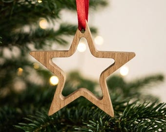 May include: A star-shaped ornament crafted from light-toned wood with a smooth finish. The center of the star is hollow, creating an open frame design. A thin, vibrant red ribbon is looped through the top point for hanging purposes.