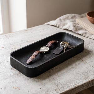 May include: A rectangular black wooden tray with rounded corners containing a wristwatch with a brown leather strap and a set of metal keys attached to a black leather fob. The tray has a visible wood grain texture throughout its surface.