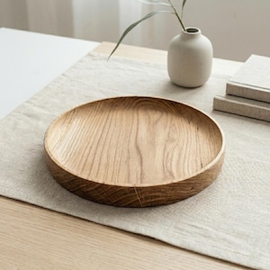 Può includere: Un vassoio rotondo in legno chiaro con bordo rialzato. Il vassoio presenta un motivo a venature naturali del legno. Un piccolo vaso bianco con una pianta e due libri sono sullo sfondo.