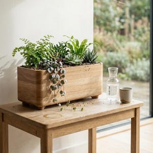 Puede incluir: Una jardinera rectangular de madera llena de varias suculentas verdes y plantas colgantes. La caja de madera de color claro está sobre una mesa de madera a juego, junto a una jarra de vidrio y una pequeña taza de cerámica.