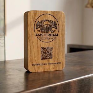 以下が含まれることがあります： 運河船と橋のレーザー刻印ロゴ、AMSTERDAM BOAT TOURSの文字、QRコード、および表面に黒で印刷されたREVIEW US ON TRIPADVISORの文字が特徴の、長方形の明るい茶色の木製看板。