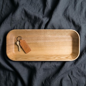 May include: A light-colored wooden tray with rounded corners. A brown leather keychain with a silver ring and two keys rests inside the tray. The tray sits on a dark gray, textured surface.