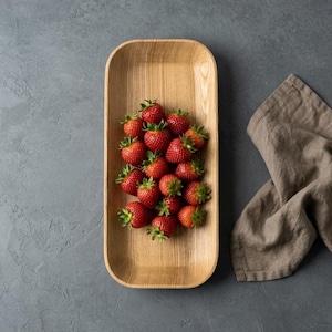 Puede incluir: Una bandeja rectangular de madera llena de fresas rojas frescas con tallos verdes. Una servilleta de lino beige está en el lado derecho de la bandeja. La bandeja y las fresas están sobre una superficie gris.