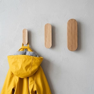 Op de afbeelding: Drie ovale wandhaken van licht hout met een natuurlijk nerfpatroon, verticaal gemonteerd. Een felgele regenjas met capuchon en grijze voering hangt aan de linkerhaak, terwijl de andere twee haken leeg aan de muur zijn bevestigd.