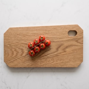 Puede incluir: Una tabla de cortar rectangular de madera con esquinas anguladas y un orificio circular para el agarre en el lado derecho. Un racimo de seis tomates pequeños de color rojo brillante con tallos verdes se sitúa en el centro de la madera.