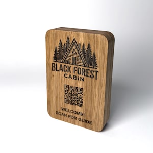 May include: A rectangular light brown wooden plaque featuring a laser-engraved graphic of a cabin surrounded by pine trees. Below the graphic, the text reads BLACK FOREST CABIN, followed by a QR code and the text WELCOME! SCAN FOR GUIDE.