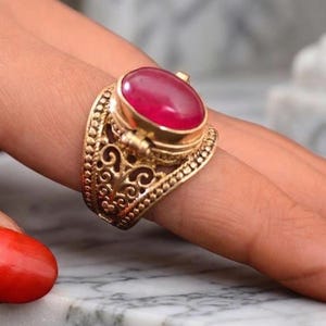 May include: A gold-colored ring with an oval, red gemstone. The ring has an ornate, filigree design around the gemstone and band. The ring is on a person's finger with red nail polish.