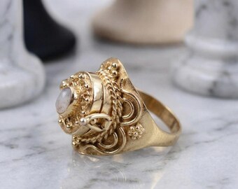 May include: A gold-colored ring with an oval, light-colored stone. The ring has an ornate design with braided and beaded details. The ring is set on a marble surface.