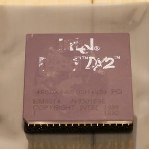 May include: A close-up of a vintage Intel 486DX2-66 processor. The processor is a rectangular shape with a dark purple top and gold pins along the bottom edge. The text "Intel" and "DX2" are visible on the top.